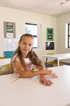 Child student sitting at white classroom table resting arms looking in pink dress mustard chairs