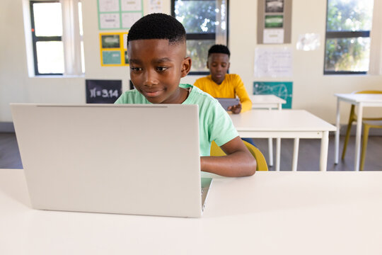 African American youth males sitting at class desks wearing green, yellow tops, using laptop tablet