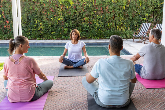 Diverse friends following senior woman practicing seated on yoga mats on brick patio beside pool