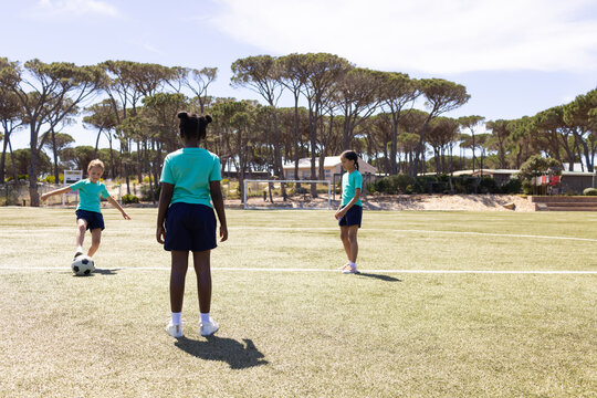 Diverse female children practicing soccer on marked field in teal shirts and navy shorts with ball