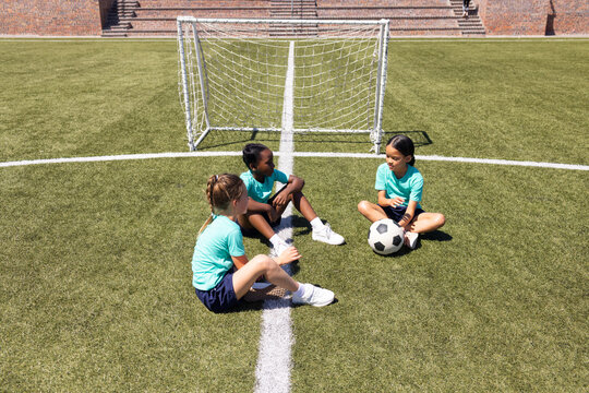 Diverse female children sitting center on turf in teal jerseys and navy shorts holding soccer ball
