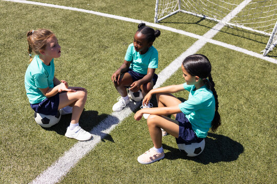 Diverse teenage female teammates sitting on soccer balls on green turf wearing matching teal shirts