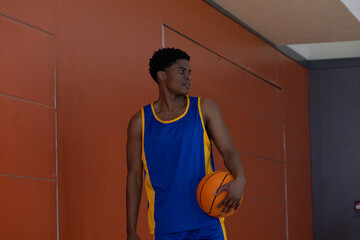 Male athlete is standing by orange paneled wall inside gym, holding basketball and looking right