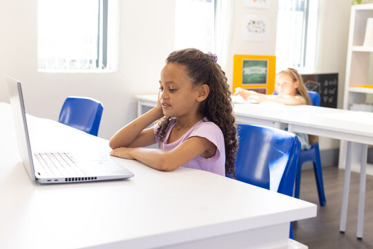 Diverse female students working on laptop at school classroom with blue chairs and bulletin board