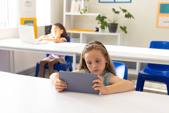 Diverse female children sitting in classroom, front child holding tablet, back child using laptop
