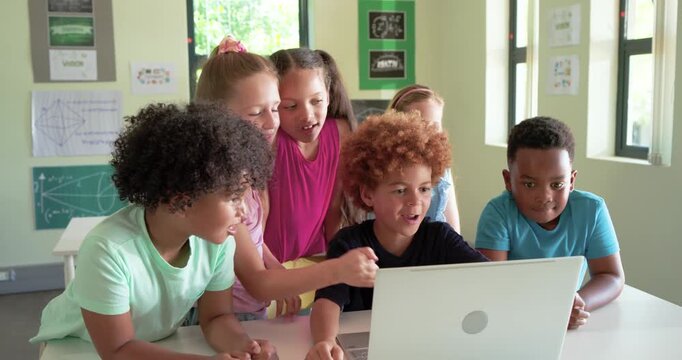 Diverse school-age children gathering around laptop in classroom, reacting to surprise on screen