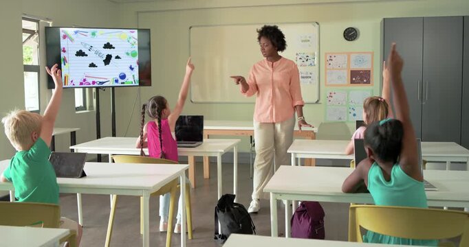 African American teacher walking in classroom scanning tablets selecting raised hand to get answers