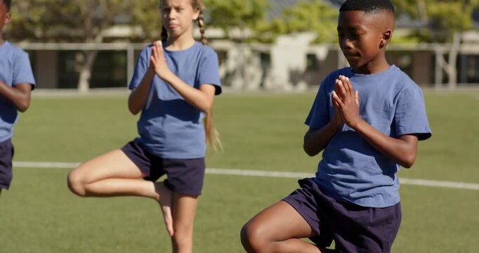 Three children wearing blue shirts holding tree pose on yoga mats at school field for PE