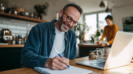 Fototapeta premium Happy middle-aged man working from home, writing in notebook with laptop on table and partner in background.