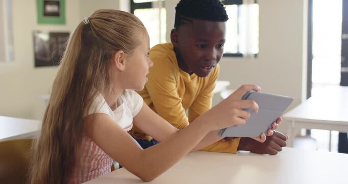 School children holding gray tablet in classroom, girl angling while boy leaning in, seeing screen