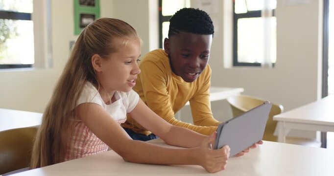 Diverse youth classmates in classroom, girl angling tablet, boy in mustard top leaning in, smiling