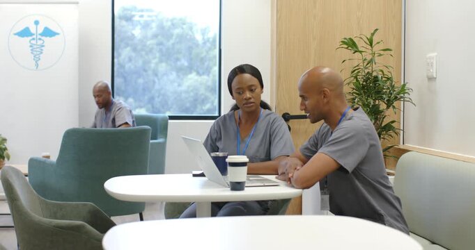 Med staff in gray scrubs in lounge, man leaning pointing, collaborating on laptop, third scrolling