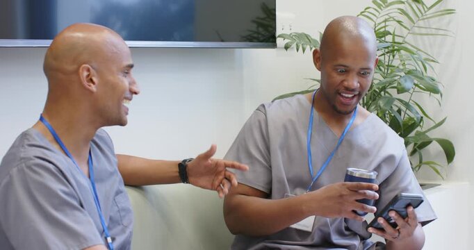 Two male medical staff in gray scrubs showing smartphone and travel mug, laughing during break
