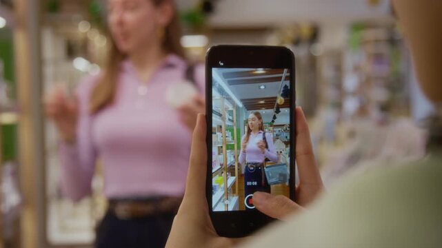 Rack focus from smartphone screen of female influencer describing nourishing hair balm on camera promoting skincare product while shooting video with friend for digital marketing at cosmetics store