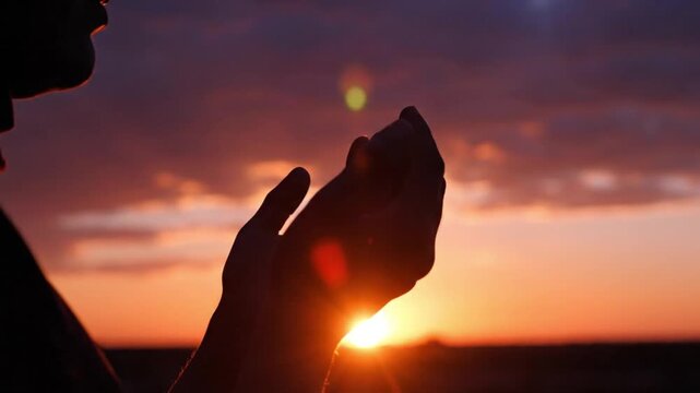A breathtaking silhouette captures a person's hands held together in a gesture of reflection or prayer against a vibrant, dramatic sunset or sunrise sky. The horizon glows with magnificent hues of ora