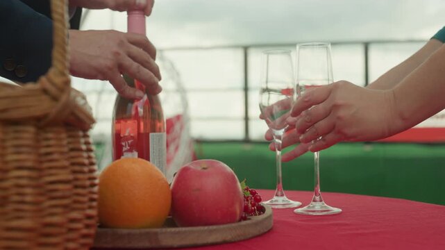 rooftop picnic with sparkling rose bottle, hands uncorking near wicker basket and two empty glasses on red tablecloth, orange and pomegranate on wooden platter, breezy skyline and playful celebratory