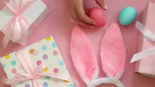 Happy Easter. Woman putting painted eggs, cute bunny ears and gift boxes on pink background, top view