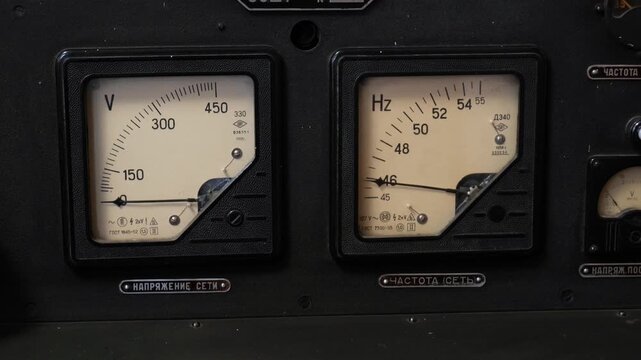 Vintage voltmeter and frequency meter with Cyrillic labels on industrial control panel, closeup panning shot of analog gauges showing 0 volts and about 46 hertz, retro electricity measurement concept