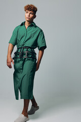 Fototapeta premium Young man with curly hair wearing green shirt dress and black belt walking barefoot on light background. Fashion model posing in casual outfit with relaxed expression and modern style.