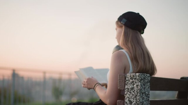 young woman reading at sunset viewpoint, backlit silhouette on bench with railing and distant skyline, headphones draped around neck, warm dusk tones and soft sky creating contemplative atmosphere