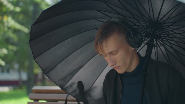 Young man under black umbrella listening with headphones to microphone while rehearsing monologue on park bench rainslick hair, pensive expression, soft natural light, green bokeh background,