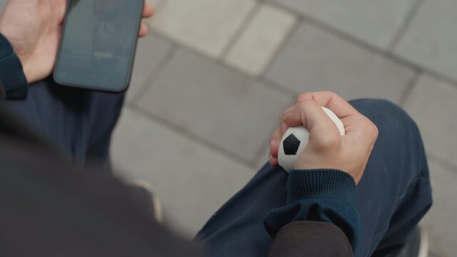 closeup hands white man holding mini soccer ball and smartphone, fingers squeezing and rotating ball to build dexterity, emphasis on texture and motion, therapeutic calm, soft bokeh urban bench