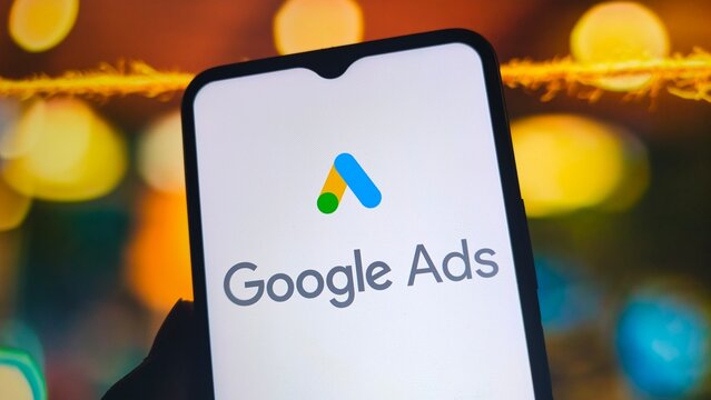 A smartphone displays the Google Ads logo on its screen with a blurred background of colorful lights.