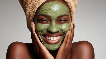 A woman with a green facial mask smiles while holding her face, showcasing a joyful skincare routine.