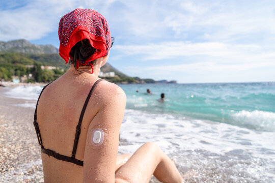 A woman wearing a red headscarf and black bikini sits on a pebbled beach facing the ocean with a visible glucose monitor on her upper arm, looking toward the water