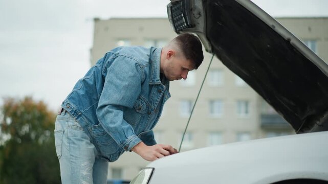 young man leaning into engine bay under raised hood, denim jacket sleeve, probing cables and oil reservoir, careful diagnostic gestures, apartment buildings in soft background, overcast light