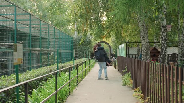 Young couple kissing on pathway under trees, intimate reunion in urban garden with birch canopy, green aviary structure to left, wooden fence and metal railing lining path, casual outfits with jackets