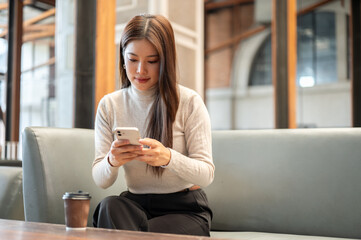 Pretty asian woman looking and using smartphone aside coffee on table while sitting on sofa in cafe.