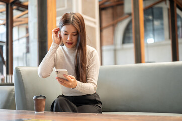 Pretty asian woman looking at phone and tucking hair aside coffee on table sitting on sofa in cafe.
