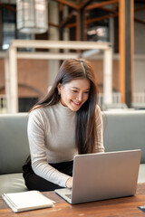 Pretty asian woman typing and looking at laptop aside book on table while sitting on sofa in a cafe.