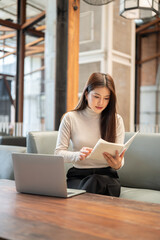 Pretty asian woman holding or reading a book aside laptop on wooden table while sits on sofa in cafe