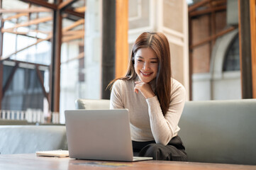 Smiling asian woman with hand under chin looking at laptop aside book on table sits on sofa in cafe.