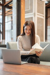 Pretty asian woman holding a book as looking at laptop on wooden table while sitting on sofa in cafe