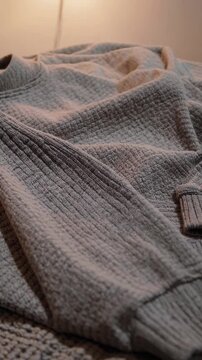 Cozy textured sweater draped on a bed, captured from a low angle. Soft lighting creates a warm, inviting atmosphere, ideal for a lifestyle video.