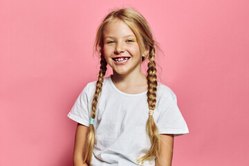 Smiling young girl with blonde hair in braids wearing a white t-shirt stands against a pink...