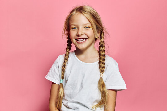 Smiling young girl with blonde hair in braids wearing a white t-shirt stands against a pink background. She shows missing teeth and looks happy and playful. Studio portrait.