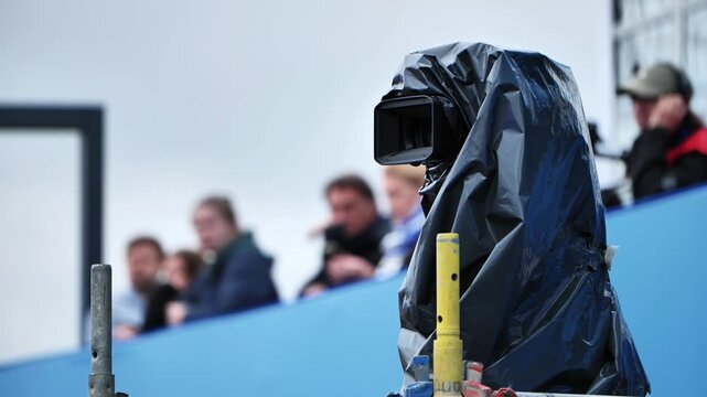 Professional video camera recording outdoor event with audience, protected from weather