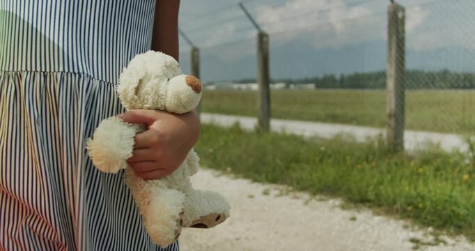 Child walks along a dirt path in a field and holds a stuffed bear close. The child looks around at the landscape. Tall grass and mountains are visible in the background.