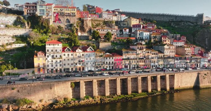 Portugal, Porto historic Ribeira district area, colorful traditional houses along Douro river waterfront, bustling road, cars, and the ancient city wall visible on the hillside. Aerial drone panorama