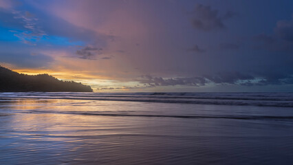 Fototapeta premium Beautiful colorful sunset over the ocean. The blue sky is highlighted with gold at the horizon. Purple and pink clouds. Waves are spreading across the beach. Reflection. A hill in the distance. 