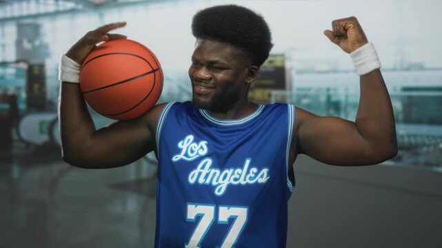 Man holding basketball on shoulder and flexing arm in airport terminal with blue los angeles jersey and white wristbands visible; confidence.
