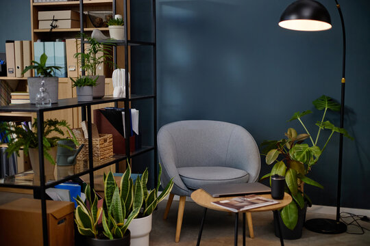 Modern office corner featuring empty armchair surrounded by green potted plants, bookshelves filled with folders and books, round table holding closed laptop and smart speaker