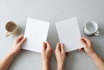 Top view of two people holding papers with grid tables for comparison on desk with mugs
