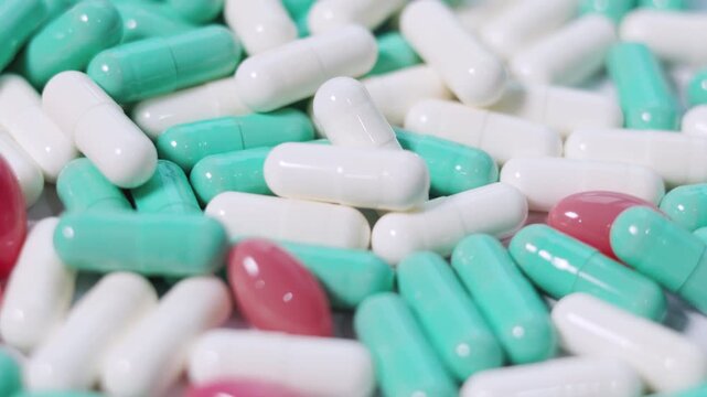 Assortment of green, white, and pink pharmaceutical capsules spins in a seamless loop. This macro footage on white surface visualizes medical treatment, drug manufacturing, and clinical research.