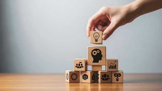 Hand building a tower with wooden blocks featuring business icons on a wooden desk with a gray background.