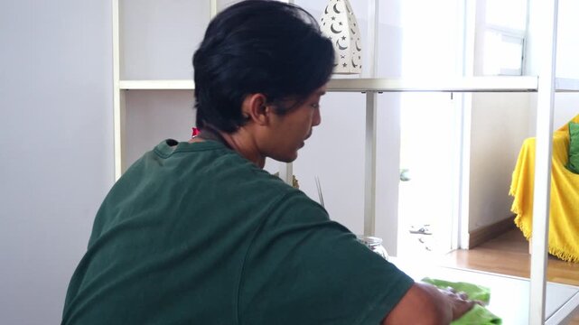 Young asian Man in apron uses colorful duster and green cloth to clean white metal shelf